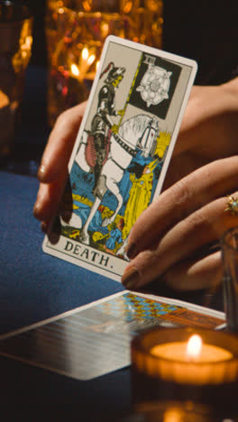 Vertical-Video-Close-Up-Of-Woman-Giving-Tarot-Card-Reading-On-Candlelit-Table-Holding-Death-Card